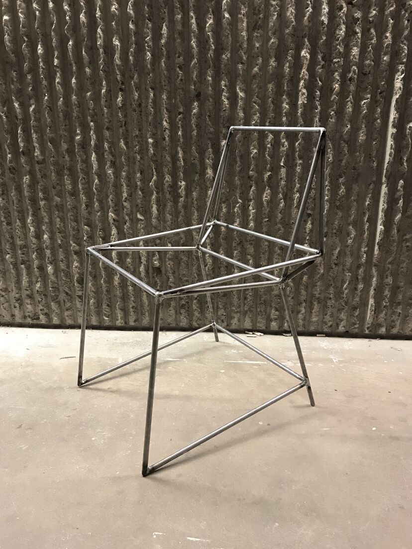 The structural frame of a chair made out of welded steel tubing sits under hash lighting in front of a corrugated concrete wall.