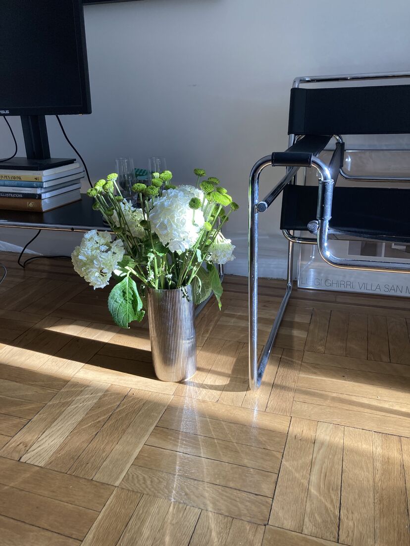 A bouquet of flowers in a silver vase on the floor of an apartment living room, reflecting rays of sunlight in a radial pattern on the parquet wood flooring.